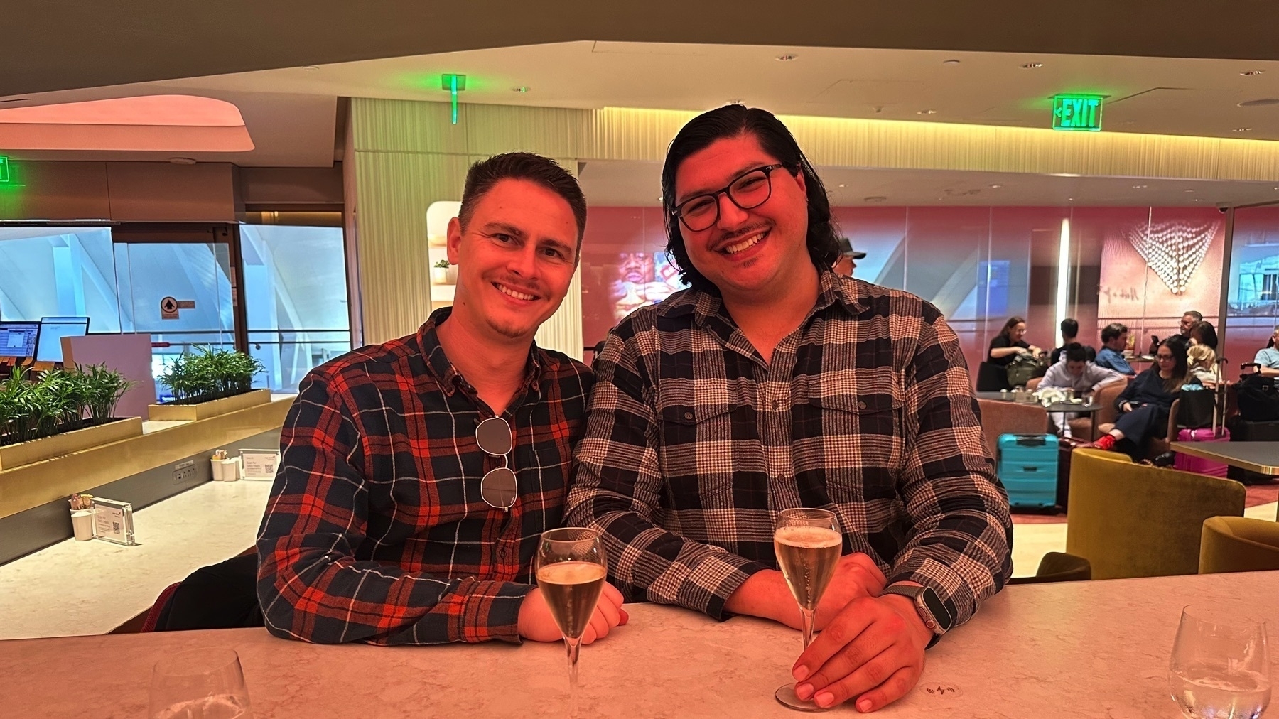 Two men smiling and holding champagne glasses while seated at a bar in an airport lounge with warm lighting and a modern design. The background shows other travelers sitting on plush chairs, some with luggage nearby.